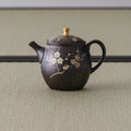 Dark bronze tokoname teapot with a rear loop handle and gold lid knob, spout to the right, floral accents near the base