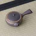 Dark clay kyusu teapot with a side handle and carved oval band, viewed from above, on a tatami mat.
