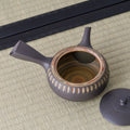 Dark clay kyusu teapot with a side handle and carved oval band, lid removed to show the swirled interior, on a tatami mat.