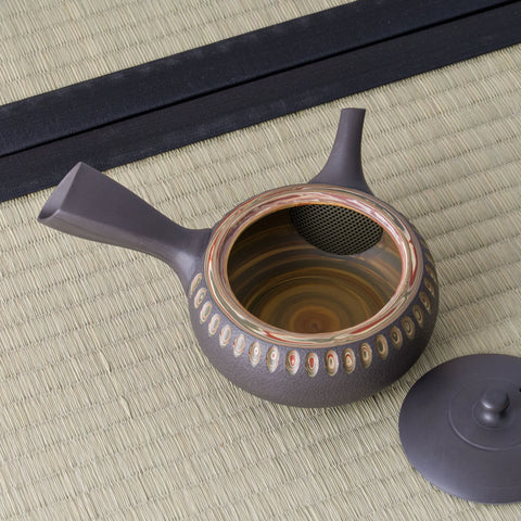 Dark clay kyusu teapot with a side handle and carved oval band, lid removed to show the swirled interior, on a tatami mat.