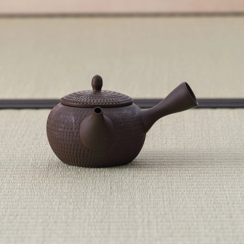 Dark brown kyusu teapot with a side handle, basket-weave carved body and lid, side view on a tatami mat.