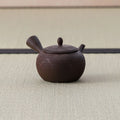 Small dark brown round kyusu with a side handle and combed body, side view on a tatami mat.