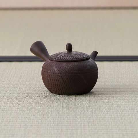 Small dark brown round kyusu with a side handle and combed body, side view on a tatami mat.