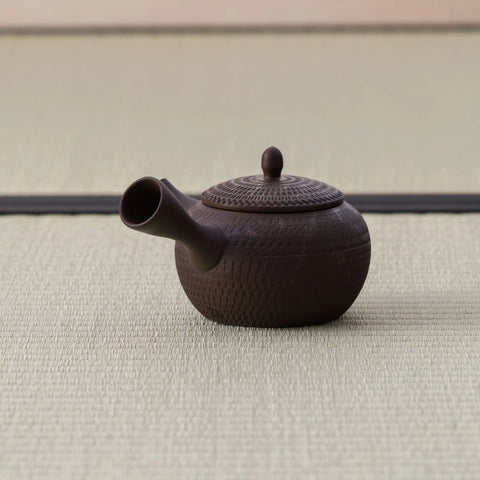Dark brown kyusu teapot with a carved body and side handle, side view on a tatami mat.