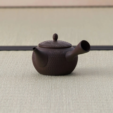 Dark brown kyusu teapot with a carved body, side handle and lid, side view on a tatami mat.