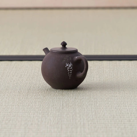 Small purple-brown kyusu with a loop handle, wisteria inlay and lid, side view on a tatami mat.