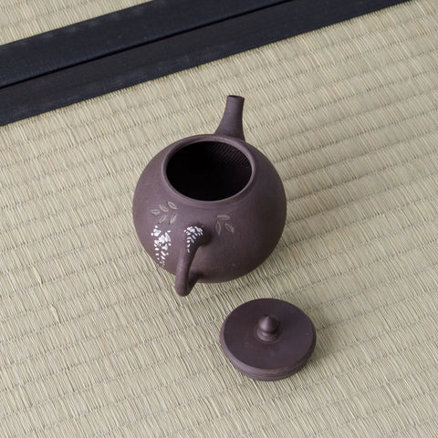 Purple-brown kyusu with a side handle, lid removed to show the mesh strainer, floral inlay, on a tatami mat.