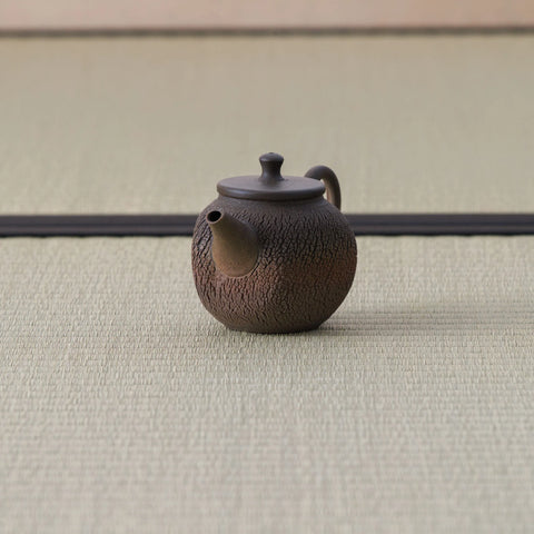 Brown wood-fired kyusu with a loop handle and textured body, side view on a tatami mat.