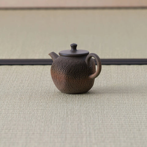 Brown wood-fired kyusu with a loop handle, textured body and lid, side view on a tatami mat.