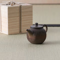 Dark wood-fired kyusu with a loop handle and lid beside a wooden box, on a tatami mat.