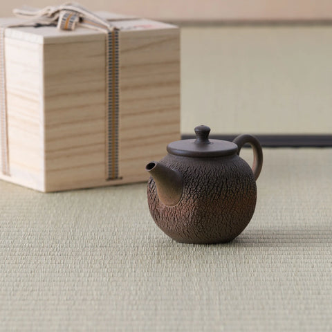 Dark wood-fired kyusu with a loop handle and lid beside a wooden box, on a tatami mat.