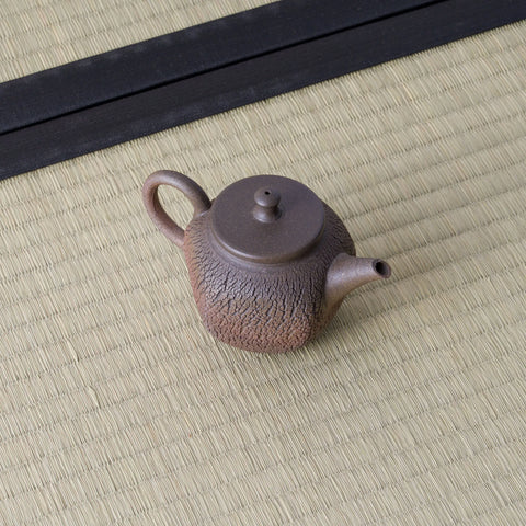 Small purple-brown kyusu with a loop handle and lid, angled view on a tatami mat.