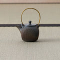 Dark wood-fired teapot with an arched brass bail handle, textured body and lid, side view.
