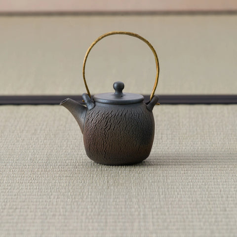 Dark wood-fired teapot with an arched brass bail handle, textured body and lid, side view.