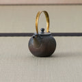 Dark wood-fired round teapot with an arched brass bail handle, side view on a tatami mat.