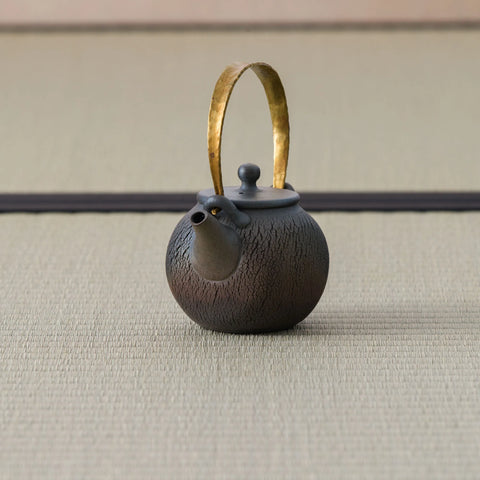 Dark wood-fired round teapot with an arched brass bail handle, side view on a tatami mat.