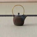 Dark wood-fired teapot with an arched brass bail handle and textured body, side view.