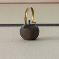 Dark wood-fired round teapot with an arched brass bail handle, side view on a tatami mat.