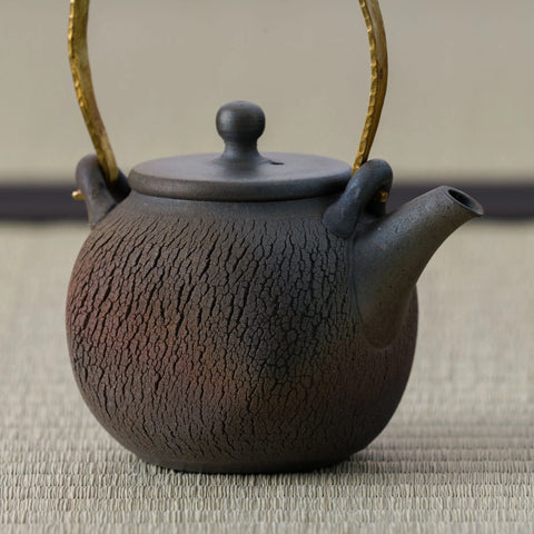 Dark wood-fired teapot with an arched brass bail handle and cracked textured body, close side view.