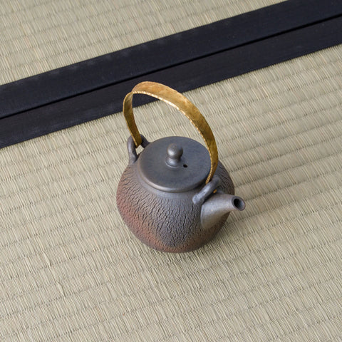 Dark wood-fired teapot with an arched brass bail handle and lid, angled view on a tatami mat.