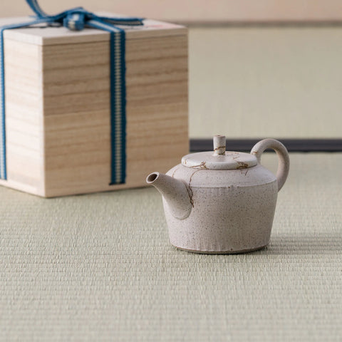 Speckled beige kyusu teapot with a loop handle and lid beside a wooden gift box, on a tatami mat.