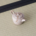 Speckled beige kyusu teapot with a loop handle and spout, three-quarter view on a tatami mat.