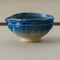 A handmade ceramic tea bowl with a blue glazed finish and a natural beige clay foot, set against a neutral textured background.
