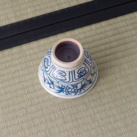 Blue-and-white porcelain bowl inverted to show its foot, decorated with a blue floral scroll, on a tatami mat.