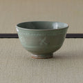 Grey-green celadon tea bowl with inlaid slip decoration, side view on a tatami mat.