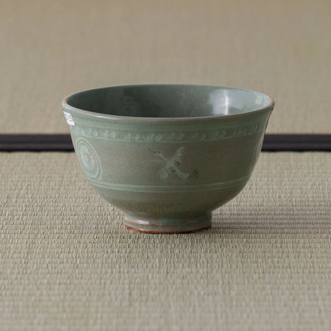 Grey-green celadon tea bowl with inlaid slip decoration, side view on a tatami mat.