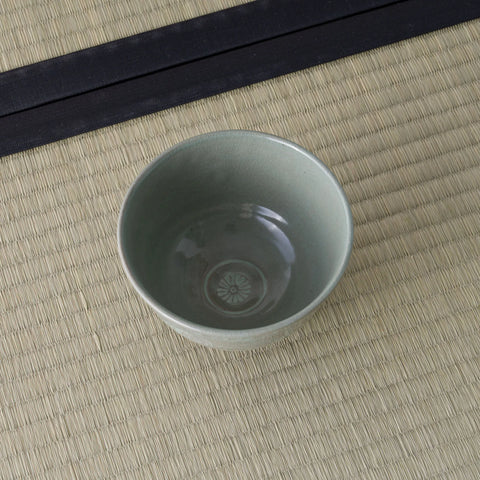 Grey-green celadon tea bowl viewed from above, showing a stamped chrysanthemum motif inside.