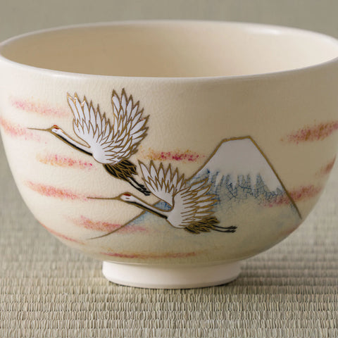 Cream tea bowl painted with cranes flying over Mount Fuji amid pink mist, close side view.