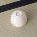 Cream tea bowl inverted to show its foot, with faint pink brushwork, on a tatami mat.
