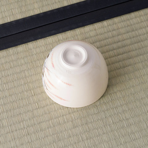 Cream tea bowl inverted to show its foot, with faint pink brushwork, on a tatami mat.