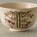 Close view of a cream tea bowl with a gold rim, painted with green bamboo and red-and-blue flowers.