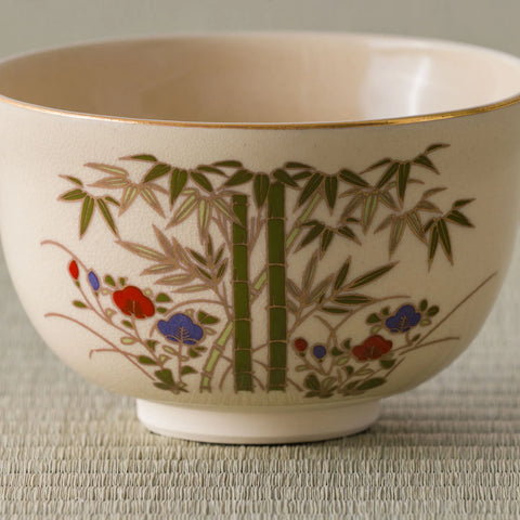 Close view of a cream tea bowl with a gold rim, painted with green bamboo and red-and-blue flowers.