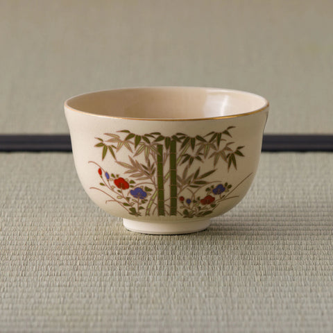 Cream tea bowl with a gold rim, painted with bamboo and red-and-blue flowers, side view.