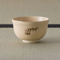 Cream tea bowl with a gold-edged rim and a small bamboo-leaf sprig, side view.