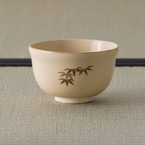 Cream tea bowl with a gold-edged rim and a small bamboo-leaf sprig, side view.