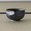 Dark metallic-glazed matcha chawan with a small pouring lip, front view on tatami