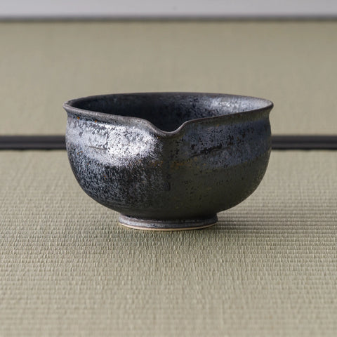 Dark metallic-glazed matcha chawan with a small pouring lip, front view on tatami