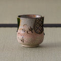 Pink crackle-glazed ceramic teacup with brown floral motif, front view on a tatami mat.