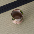 Small pink crackle-glazed ceramic teacup with a brown floral motif and green-glazed rim, on a tatami mat.