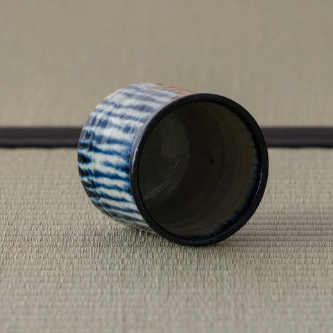 Blue-and-white striped teacup tipped on its side showing the dark interior, on a tatami mat.