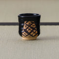 Back view of a black-glazed yunomi showing a crosshatch lattice pattern painted on the bare clay window.