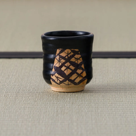 Back view of a black-glazed yunomi showing a crosshatch lattice pattern painted on the bare clay window.