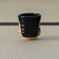 Ribbed black yunomi angled to show parts of both painted clay panels, on a tatami mat.