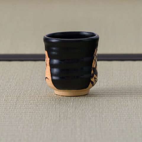 Ribbed black yunomi angled to show parts of both painted clay panels, on a tatami mat.