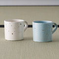 Two speckled mugs shown together for comparison, one in pale pink and one in blue-grey