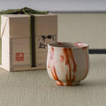 Cream wood-fired yunomi with orange-red flame markings shown with its wooden storage box on tatami